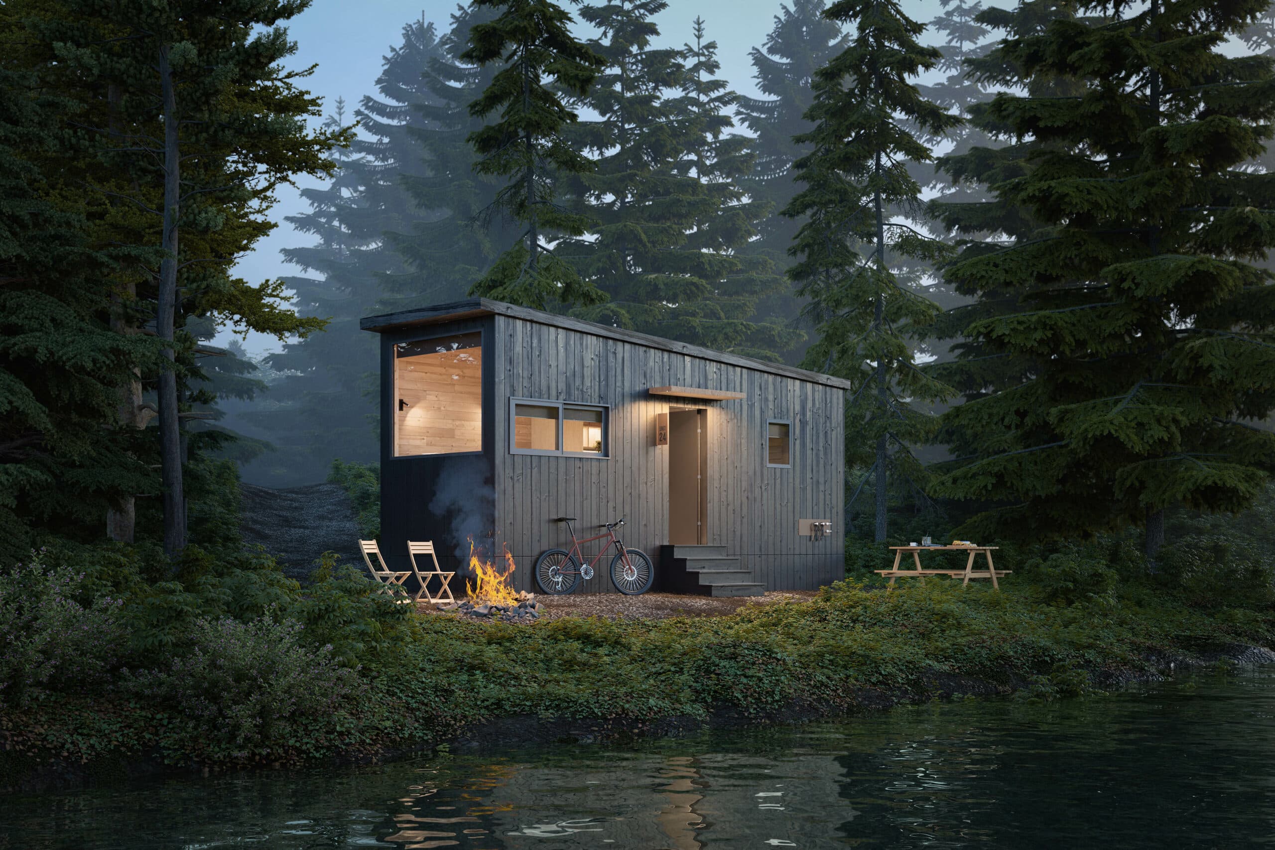 A modern tiny home with dark wood siding is nestled in a lush forest by the edge of a calm lake. The home features large windows that reveal a warmly lit wooden interior. Outside, a campfire burns brightly, surrounded by folding chairs and a parked bicycle. A picnic table is visible to the side, blending into the serene natural setting of tall evergreen trees and dense foliage.