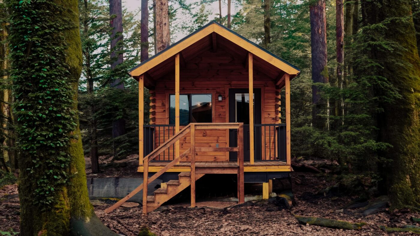 Rustic Aspen park model tiny home with a covered front porch in a wooded setting in Maine.