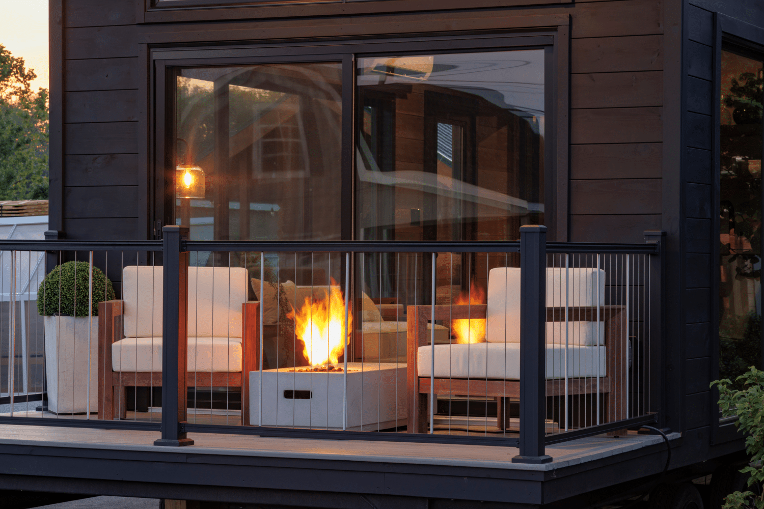 Close-up view of a modern tiny home's front porch featuring two wooden chairs with white cushions and a sleek rectangular fire pit. The porch is enclosed with black metal railings and accented by a potted plant, creating a cozy and elegant outdoor seating area. Reflections in the large glass doors showcase a warm, inviting interior and soft evening lighting.