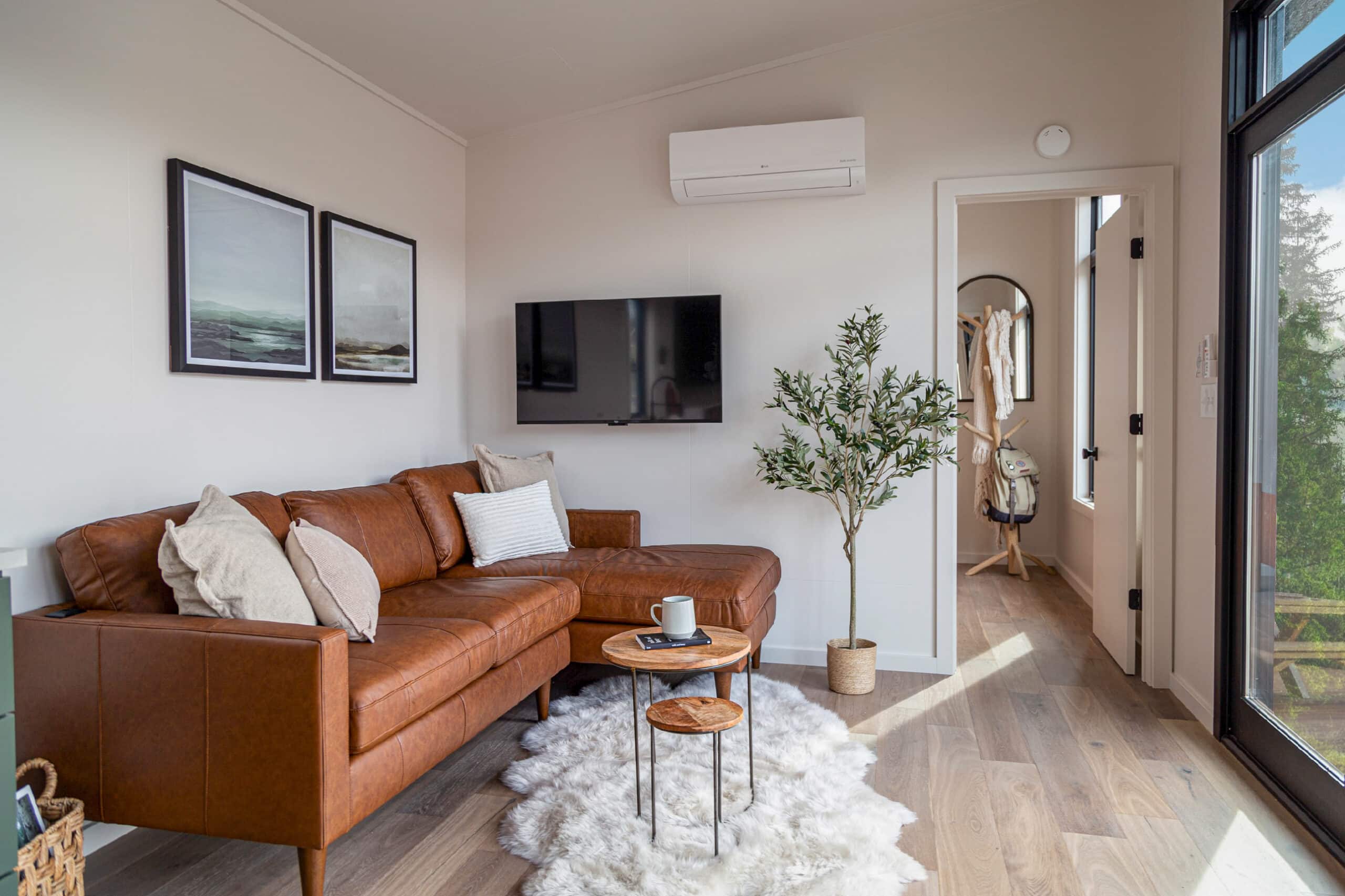 A cozy and modern tiny home interior featuring a stylish brown leather sectional sofa adorned with neutral-toned cushions, set against a light-colored wall with two framed landscape prints. A sleek wall-mounted TV is positioned above, accompanied by a mini-split air conditioner for climate control. A soft, fluffy white rug adds warmth to the wood laminate flooring, while a small set of nesting tables holds a coffee mug and book. A potted plant adds a touch of greenery, and the bright natural light streaming through a large glass door enhances the inviting ambiance. A hallway leads to a coat rack with accessories neatly organized, completing the space's functional yet elegant design.
