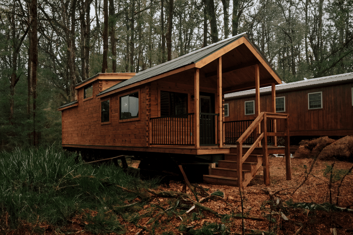 A rustic park model home with rich wooden siding and a pitched metal roof sits elevated on a wooded lot. The front porch, framed by sturdy wooden posts and railings, adds charm and functionality, complete with a staircase leading to the entrance. Large windows provide natural light, blending the home's cozy interior with the serene forest surroundings. In the background, another similarly styled cabin is visible, enhancing the community feel of the setting while maintaining its connection to nature. The lush greenery and tall trees create a tranquil and secluded atmosphere.