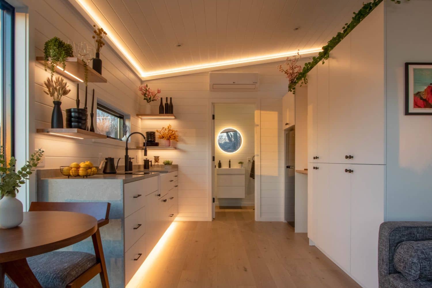 Modern tiny home interior featuring a sleek kitchen with open shelving, warm accent lighting, and a minimalist bathroom with a circular mirror