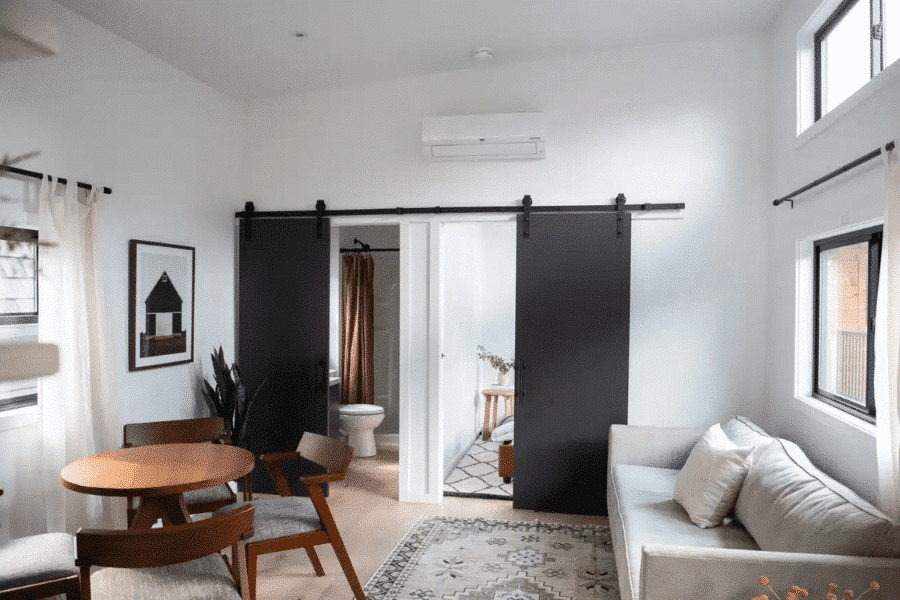 Stylish tiny home interior featuring a cozy living area with a sofa, a small dining table with chairs, and black sliding barn doors leading to a bathroom and bedroom