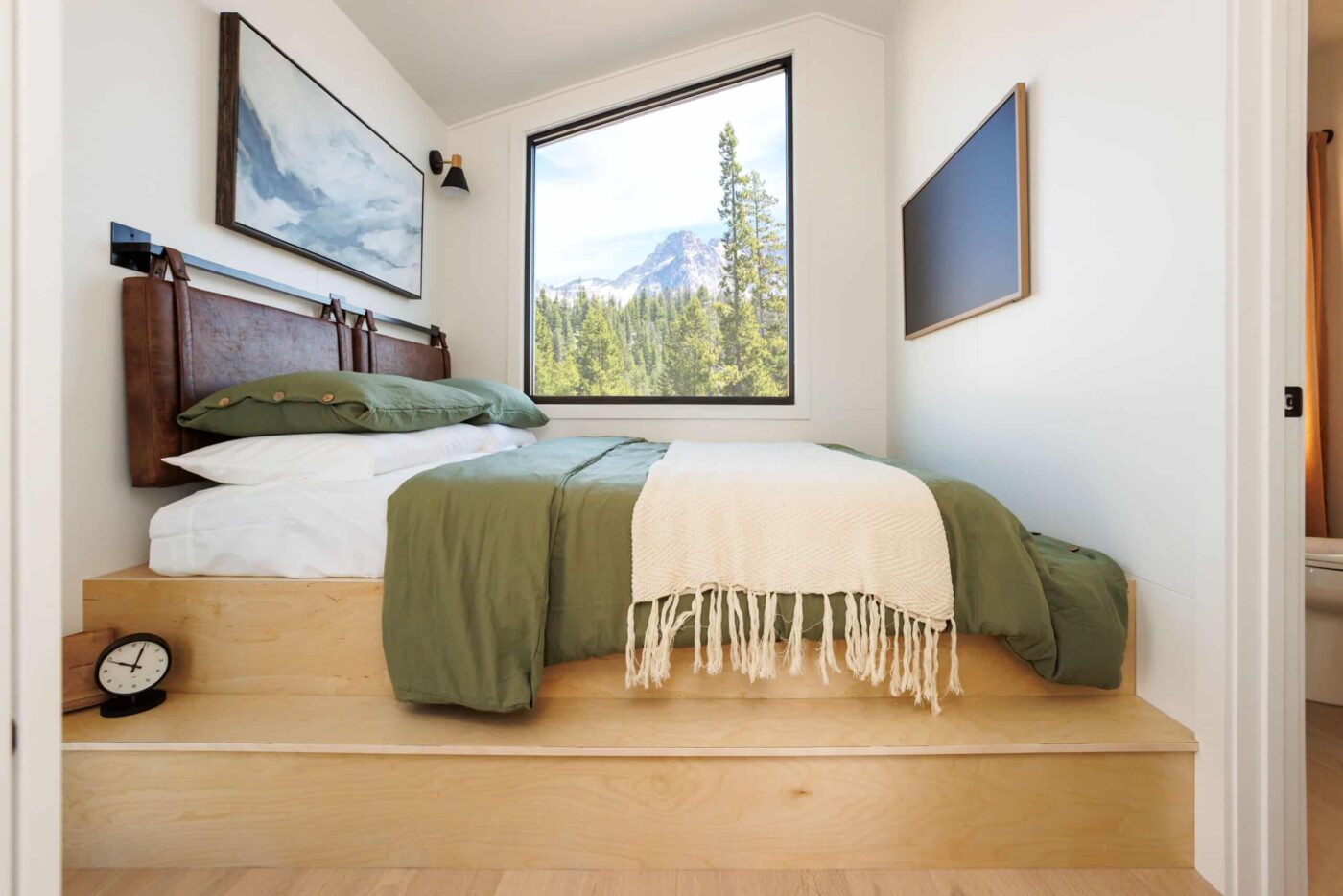 Cozy bedroom inside an A-Frame park model with a raised wood platform bed, green bedding, and large window overlooking pine trees and mountains.
